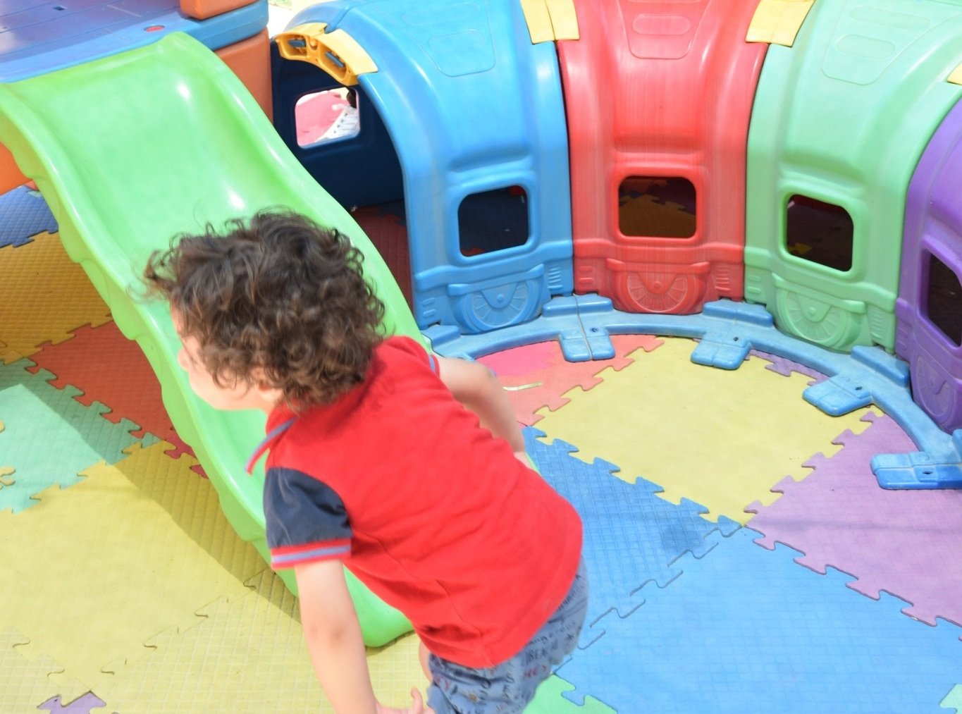A young child playing on a green slide of our toy bridge in a colorful playground environment. The child is smiling playfully and gesturing with their hands. Surrounding the child are bright yellow and orange chairs, and the walls are decorated with cartoon characters.